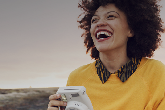 Lachende Frau mit Afro-Haar und gelbem Sweatshirt hält eine weiße Sofortbildkamera in einer weiten Landschaft