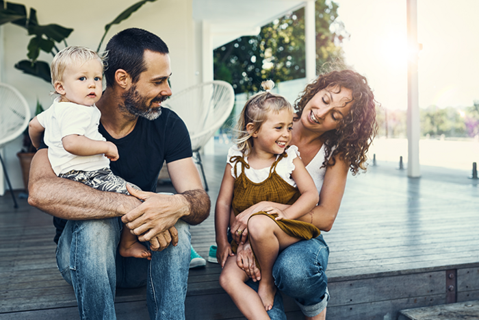 Familie mit zwei Kleinkindern sitzt lachend auf einer sonnigen Holzterrasse vor einem modernen Haus